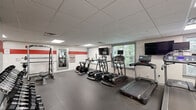 Located at 1 Parker Ave in the Roxborough neighborhood of Philadelphia, PA, this fitness center offers a comprehensive workout facility within a single, open-plan floor. The space is designed with functionality in mind, featuring durable rubber flooring throughout that provides both safety and comfort during exercise.

The cardio zone is well-appointed with multiple treadmills, elliptical machines, and stationary cycling bikes, each equipped with individual monitors for entertainment. Multiple flat-screen TVs are mounted along the walls, and windows along one side of the room allow natural light to filter in while offering views of the surrounding greenery.

The strength training area includes a multi-functional cable machine, a power rack with an adjustable bench, a dumbbell rack, and a medicine ball station — catering to a wide range of workout routines. Full-length mirrors are strategically placed to support form monitoring during exercises.

The interior is finished with a clean white color scheme accented by bold red horizontal stripes along the walls, complemented by motivational typographic wall decals that energize the atmosphere. Recessed and panel lighting in the drop ceiling ensures the space is uniformly and brightly lit. The overall ambiance is modern, clean, and purpose-driven, making this fitness center a well-rounded facility for residents or members seeking a complete workout experience.