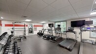 Located at 1 Parker Ave in the Roxborough neighborhood of Philadelphia, PA, this fitness center offers a comprehensive workout facility within a single, open-plan floor. The space is designed with functionality in mind, featuring durable rubber flooring throughout that provides both safety and comfort during exercise.

The cardio zone is well-appointed with multiple treadmills, elliptical machines, and stationary cycling bikes, each equipped with individual monitors for entertainment. Multiple flat-screen TVs are mounted along the walls, and windows along one side of the room allow natural light to filter in while offering views of the surrounding greenery.

The strength training area includes a multi-functional cable machine, a power rack with an adjustable bench, a dumbbell rack, and a medicine ball station — catering to a wide range of workout routines. Full-length mirrors are strategically placed to support form monitoring during exercises.

The interior is finished with a clean white color scheme accented by bold red horizontal stripes along the walls, complemented by motivational typographic wall decals that energize the atmosphere. Recessed and panel lighting in the drop ceiling ensures the space is uniformly and brightly lit. The overall ambiance is modern, clean, and purpose-driven, making this fitness center a well-rounded facility for residents or members seeking a complete workout experience.