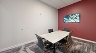 Located at 100 Pine St W in Wilson, NC, this single-room private office is a well-appointed, self-contained workspace designed for focused meetings and professional collaboration. The room is accessed through a sleek glass-paneled door that provides visual connectivity to the surrounding area while maintaining privacy, and the white tile flooring at the entry transitions seamlessly into patterned, textured carpet within the office.

The interior is anchored by a striking marble-top conference table surrounded by cushioned black executive chairs with gold-toned legs, combining elegance with functionality. Seating accommodates a small group, making it ideal for intimate meetings or collaborative sessions. A bold deep-red accent wall adds a dynamic visual contrast to the otherwise neutral, light-gray palette of the remaining walls, lending the space a sense of energy and character.

Two framed art pieces—one an abstract canvas in warm yellows, reds, and blues, the other a wall-mounted display screen—add creative dimension to the office. The ceiling features a standard drop-tile grid with recessed lighting, ensuring consistent, even illumination throughout. A glass entry wall further opens the space visually, maintaining a bright and airy feel. Overall, the office presents a refined, contemporary environment well-suited for professional use.