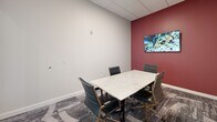 Located at 100 Pine St W in Wilson, NC, this single-room private office is a well-appointed, self-contained workspace designed for focused meetings and professional collaboration. The room is accessed through a sleek glass-paneled door that provides visual connectivity to the surrounding area while maintaining privacy, and the white tile flooring at the entry transitions seamlessly into patterned, textured carpet within the office.

The interior is anchored by a striking marble-top conference table surrounded by cushioned black executive chairs with gold-toned legs, combining elegance with functionality. Seating accommodates a small group, making it ideal for intimate meetings or collaborative sessions. A bold deep-red accent wall adds a dynamic visual contrast to the otherwise neutral, light-gray palette of the remaining walls, lending the space a sense of energy and character.

Two framed art pieces—one an abstract canvas in warm yellows, reds, and blues, the other a wall-mounted display screen—add creative dimension to the office. The ceiling features a standard drop-tile grid with recessed lighting, ensuring consistent, even illumination throughout. A glass entry wall further opens the space visually, maintaining a bright and airy feel. Overall, the office presents a refined, contemporary environment well-suited for professional use.