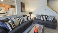 This two-floor space offers a comfortable and inviting living environment with a clean, modern aesthetic. The living area serves as a central gathering space, featuring a dark charcoal sofa and matching armchair dressed with a mix of patterned and striped throw pillows in black, white, and neutral tones, creating a stylish yet relaxed atmosphere. A glass-top coffee table anchors the seating arrangement, adding a touch of contemporary elegance while maintaining an open, airy feel underfoot with soft carpeting. Ambient lighting is provided by a classic table lamp, contributing to the warm, cozy ambiance of the room. A framed art piece depicting a rainy street scene adds a subtle artistic focal point to the neutral-toned walls. The open layout between areas is suggested by a partial wall cutout, hinting at a connected adjacent space — potentially a kitchen or dining area — which enhances the sense of flow throughout the main level. Spanning two floors, this space provides distinct living zones, offering both communal and private areas suited for comfortable everyday living.