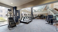 This expansive fitness center is designed to deliver a premium workout experience across a thoughtfully organized, single-level layout that benefits from soaring double-height ceilings and an open mezzanine above. The main floor is anchored by a dedicated cardio zone featuring a row of treadmills, stair climbers, ellipticals, and stationary bikes, each equipped with individual display screens. Large windows along the perimeter allow natural light to fill the space, creating an energizing atmosphere. Adjacent to the cardio area, a comprehensive strength training zone houses an extensive selection of Matrix-branded resistance machines, weight benches, and a full dumbbell rack, all reflected in floor-to-ceiling mirrors that amplify the sense of space. A raised platform area, accessed by wood-accented stairs with glass and metal railings, provides a distinct section for additional equipment and functional training. Overhead, wall-mounted flat-screen televisions are positioned throughout for convenient viewing during workouts. The flooring throughout features patterned carpet tile in a neutral blue-grey palette, complementing the facility's modern navy and warm beige color scheme. Recessed and ambient lighting enhances visibility across all zones, while ceiling fans provide additional comfort. A separate enclosed room offers a more private strength training environment, also finished with mirrored walls and consistent design elements.