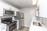 This centrally located two-bedroom unit is laid out on a single floor, offering a practical and well-organized living space. The living area is generously sized, featuring updated wood-look hard-surface flooring in a cool gray tone that extends throughout the main living spaces. Neutral warm-gray walls and a textured ceiling create a clean, understated backdrop. Natural light enters through a large window fitted with blinds, while a sliding glass door with vertical blinds provides outdoor access and additional brightness. A wall-mounted air conditioning unit and baseboard heating ensure year-round comfort.

The kitchen is equipped with stainless steel appliances, including a refrigerator, electric range, over-the-range microwave, and dishwasher. White shaker-style cabinetry and white subway tile backsplash complement the light countertops, while wood-look flooring continues throughout the space.

Both bedrooms feature plush carpeting, neutral walls, and individual windows that bring in natural light and outdoor views of surrounding greenery. Baseboard heaters are present in each room.

The bathroom is finished with wood-look flooring, a white vanity with shaker-style cabinetry, a medicine cabinet mirror, and a full tub-shower combination, all presented in a clean, bright palette.