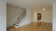 This two-story residence offers a well-organized layout designed for comfortable everyday living. The main level is anchored by an open living area finished with warm, light-toned hardwood floors that extend seamlessly throughout the space, lending a sense of continuity and warmth. A staircase with white-painted risers and a classic wrought iron railing with decorative detailing serves as a subtle focal point, connecting the two floors with understated elegance. The neutral white walls throughout the main level create a bright, airy atmosphere that enhances the sense of space. Toward the rear of the main floor, a dedicated kitchen is visible, featuring warm wood cabinetry, dark appliances, and ample counter space — a practical and well-appointed area for cooking and meal preparation. A hallway corridor provides clear circulation between the living areas and the kitchen, contributing to the home's efficient floor plan. The upper level, accessible via the staircase, offers additional living or sleeping quarters. Overall, the space presents a clean, move-in-ready aesthetic with classic design choices, durable finishes, and a cohesive flow between rooms that makes it both functional and inviting.