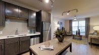 This single-floor residence showcases a thoughtfully designed open-concept layout that seamlessly connects the kitchen, dining, and living areas into one cohesive living space. The kitchen is outfitted with rich, dark espresso cabinetry that extends to the ceiling, offering generous storage alongside sleek stainless steel appliances, including a full-size refrigerator and dishwasher. Light-toned countertops provide a striking contrast to the dark cabinetry, while a built-in sink adds to the kitchen's functional design. Overhead, a track lighting system illuminates the space with a modern, industrial flair. The adjacent dining area features a sturdy wooden table, ideal for everyday meals or entertaining guests. Flowing naturally from the dining space, the living room is bathed in natural light streaming through large windows dressed with elegant curtains. Warm hardwood-style flooring runs throughout, unifying the open floor plan. The living area offers a relaxed and inviting ambiance, complemented by tasteful décor and soft furnishings. Overall, this residence strikes a refined balance between contemporary style and everyday comfort.