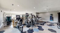 Located in Austin, TX, this fitness center offers a thoughtfully designed, single-floor exercise space that accommodates a comprehensive range of workout options. The room is divided into two distinct zones: a cardio area and a strength training area, providing a well-rounded experience for users of all fitness levels.

The cardio zone features a stylish hexagonal-patterned carpet in shades of blue and gray, lending a modern aesthetic to the space. It is equipped with multiple treadmills, elliptical trainers, and stationary bikes, each outfitted with digital display screens. Wall-to-wall mirrors amplify the sense of space, while recessed lighting provides bright, even illumination throughout.

The strength training area transitions to light wood-look flooring, creating a clean and energizing atmosphere. Large windows along the walls invite ample natural light and offer views of the surrounding Austin landscape. This zone includes a multi-functional cable machine, weight benches, a full rack of dumbbells, and additional selectorized strength machines, catering to a wide variety of training routines.

A digital countdown clock is mounted on the wall for interval training, and a foam roller and accessory storage area add practical functionality. The neutral gray walls, recessed ceiling lights, and contemporary equipment collectively contribute to a clean, professional, and motivating environment well-suited for daily fitness activities.