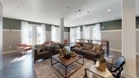 This well-appointed single-floor amenity space blends contemporary design with comfortable, functional areas suited for socializing and recreation. The main lounge area is generously sized, featuring warm hardwood-style flooring, two-tone walls with a rich green upper accent and neutral lower paneling, and recessed lighting throughout. Plush seating arrangements, including deep sofas and accent chairs, are anchored by a wood-top coffee table, creating an inviting gathering space. Large windows dressed with sheer white drapes allow abundant natural light to fill the room. A dedicated seating alcove off the main lounge offers a quieter retreat with modern white armchairs and small side tables. The game area is a standout feature, showcasing exposed wood beam ceiling details, a sleek wet bar with light wood cabinetry, white quartz countertops, subway tile backsplash, and dark upper cabinets with stylish globe sconces. A wall-mounted billiard cue rack signals the space's recreational purpose. Multiple sets of French doors and floor-to-ceiling windows open directly onto an outdoor deck, offering views of a community pool area and providing seamless indoor-outdoor connectivity. The overall ambiance is modern, warm, and social — thoughtfully designed for community living.