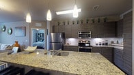 This single-floor residence showcases a cohesive modern design throughout its thoughtfully arranged layout. The kitchen serves as a central highlight, featuring sleek dark wood-grain cabinetry extending to the ceiling, stainless steel appliances including a range, microwave, and refrigerator, and granite countertops that continue across a generously sized island with an integrated sink. White subway tile backsplash and under-cabinet lighting add a polished, contemporary finish, while rich dark hardwood flooring grounds the space.

The living area offers an inviting atmosphere with dark hardwood floors, large windows dressed in sheer teal curtains and slatted blinds, a ceiling fan, and direct access to an outdoor area, allowing ample natural light and outdoor views.

The bedrooms feature plush carpet, multiple windows with outdoor views, and ceiling fans, creating comfortable, well-lit retreats. One bedroom includes walk-in closet access directly from the bathroom.

The bathrooms are finished with large-format tile flooring, granite-topped dark wood vanities, and well-organized walk-in closets visible beyond. One bathroom includes a soaking tub with shower combination. The overall ambiance is clean, modern, and cohesive throughout.
