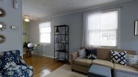 This single-floor space offers a warm and inviting atmosphere defined by classic design details and thoughtful layout. The living area features rich hardwood flooring that anchors the room with a sense of warmth, complemented by soft blue-gray walls that lend a calm, airy feel throughout. Crown molding adds a refined architectural touch, while a textured ceiling provides subtle character overhead. Multiple windows dressed with sheer white curtains and valances allow generous natural light to fill the room, enhancing the open and comfortable ambiance. A plush area rug anchors the seating area, where a tan sofa accented with decorative throw pillows in navy and blue tones creates an inviting focal point. A sleek black open-frame bookshelf serves both as storage and a display feature, adding an industrial contrast to the otherwise soft, neutral palette. A glass-top dining or accent table with acrylic chairs introduces a modern, airy element to the space. The overall design blends casual comfort with understated elegance, making this a functional and visually cohesive living environment well-suited for everyday living and relaxed entertaining.