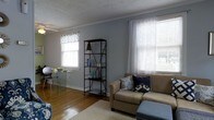 This single-floor space offers a warm and inviting atmosphere defined by classic design details and thoughtful layout. The living area features rich hardwood flooring that anchors the room with a sense of warmth, complemented by soft blue-gray walls that lend a calm, airy feel throughout. Crown molding adds a refined architectural touch, while a textured ceiling provides subtle character overhead. Multiple windows dressed with sheer white curtains and valances allow generous natural light to fill the room, enhancing the open and comfortable ambiance. A plush area rug anchors the seating area, where a tan sofa accented with decorative throw pillows in navy and blue tones creates an inviting focal point. A sleek black open-frame bookshelf serves both as storage and a display feature, adding an industrial contrast to the otherwise soft, neutral palette. A glass-top dining or accent table with acrylic chairs introduces a modern, airy element to the space. The overall design blends casual comfort with understated elegance, making this a functional and visually cohesive living environment well-suited for everyday living and relaxed entertaining.