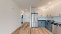 This thoughtfully designed single-floor unit offers a clean, contemporary aesthetic defined by crisp white walls, light-toned luxury vinyl plank flooring, and an abundance of natural light. The open-concept layout connects the kitchen and living area seamlessly, creating a spacious and functional flow. The kitchen is a standout feature, showcasing two-tone cabinetry with soft blue-gray lower cabinets and white upper cabinets, a light blue subway tile backsplash, dark countertops, and a full suite of stainless steel appliances including a French door refrigerator, electric range, over-the-range microwave, and dishwasher. Recessed lighting overhead adds a polished finish to the space. The living area is generously proportioned, with large windows fitted with roller shades that invite natural light and offer open outdoor views. A wall-mounted HVAC unit ensures year-round comfort. The entryway features an electrical panel access point and leads directly to the main bathroom, which includes large-format tile, a white vanity with an undermount sink, and a glass-enclosed walk-in shower with grab bars. A second bathroom offers similar clean finishes with a toilet and vanity. The bedrooms are bright and well-sized, each benefiting from multiple windows with exterior views and hard-surface flooring throughout.