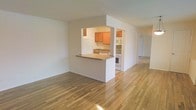 This single-floor space showcases a clean, open-concept layout with warm hardwood floors extending throughout the main living area, reflecting natural light and adding a sense of warmth and continuity. The kitchen is thoughtfully designed as a semi-open pass-through, framed by a half-wall counter that separates it from the living area while maintaining visual connectivity. Honey-toned wood cabinetry lines the kitchen walls, complemented by white appliances including a range, microwave, and refrigerator, alongside a functional sink set into a laminate countertop. The pass-through counter offers additional prep or casual dining space. The surrounding living area is notably open and airy, with smooth white walls and a neutral palette that provides a versatile backdrop for a variety of interior styles. A hallway visible toward the rear of the space suggests additional rooms beyond the main living area. The overall ambiance is clean and understated, with the polished hardwood floors serving as a standout feature that anchors the space with warmth and character.