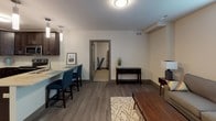 This single-floor unit offers a thoughtfully designed open-concept layout that seamlessly connects the kitchen, dining, and living areas. The kitchen is outfitted with dark espresso cabinetry, granite countertops, stainless steel appliances, and a peninsula with bar seating — making it both functional and visually striking. Durable wood-style flooring extends throughout the main living areas, complementing the neutral color palette and contemporary finishes.

The living room is a comfortable, well-proportioned space featuring a window that allows natural light to filter in, creating a warm and inviting atmosphere. A console table near the entry adds a practical transitional element between the living and kitchen areas.

Two bedrooms provide restful retreats, each finished with plush carpeting. One bedroom features dual windows and a ceiling fan with integrated lighting, enhancing ventilation and natural light. The second bedroom is equally comfortable, with soft tones and easy access to storage.

The full bathroom is finished with tile flooring, a dark vanity with granite countertop, a large mirror, and a tub-shower combination. An additional versatile bonus room with neutral carpet offers flexible use as a home office or den.