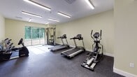 This single-floor amenity space in North Smithfield, Rhode Island, is thoughtfully designed to offer a range of communal functions within a cohesive, modern layout. The fitness center is a standout feature, equipped with treadmills, elliptical machines, a multi-station cable system, and a dedicated free-weight area with a full dumbbell rack and workout bench. Rubber flooring throughout the gym provides durability and safety, while large windows invite natural light and offer views of the surrounding greenery. A sitting area adjacent to the fitness space features two tufted navy blue accent chairs centered around a round wooden coffee table atop a patterned area rug, creating an intimate and comfortable retreat. The lounge area is anchored by a rich leather sofa set against a striking wood-planked accent wall, with French doors opening to the outdoors. The dining area showcases dark hardwood-style flooring, a bold teal accent wall, pendant lighting, and a wet bar with dark cabinetry, complemented by large windows framing wooded views. Dual water fountains are conveniently recessed into a hallway niche, adding a practical amenity to this well-appointed space.