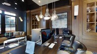 Located in the heart of West Chester, PA, this single-floor space at 201 E Gay Street offers a polished and professional environment designed for productivity and comfort. The interior exudes a refined, upscale aesthetic anchored by rich dark wood paneling, deep-toned walls, and warm hardwood surfaces that create an atmosphere of focused elegance. A generously sized communal work desk runs along the center of the room, outfitted with multiple iMac workstations and equipped with a shared printer, making it a fully functional business center. Overhead, a cluster of glass pendant lights with metallic accents provides focused task lighting while contributing to the room's contemporary character. Built-in cabinetry with display shelving lines one wall, adding both storage and visual depth to the space. A large-scale abstract artwork mounted in a wood-framed panel serves as a striking focal point, balancing the room's darker tones with subtle texture and color. Leather seating adds a touch of executive comfort. The overall ambiance is one of sophisticated utility — professional in purpose, yet thoughtfully appointed in design — making it an ideal setting for work and business-related tasks.