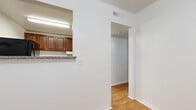 This single-floor unit offers a practical and straightforward layout with distinct zones for living, cooking, and bathing. The kitchen is equipped with rich, dark wood cabinetry extending to the upper walls, providing ample storage. Granite-patterned countertops line the workspace, complemented by a stainless steel sink, a black gas range, a microwave, and a full-size black refrigerator. Light ceramic tile flooring runs throughout the kitchen area. A pass-through window opening connects the kitchen to the adjacent hallway, enhancing the sense of openness between spaces.

The bedrooms feature dark hardwood-style flooring and are each fitted with ceiling fans with integrated lighting. One bedroom benefits from a window offering a view of surrounding greenery, bringing in natural light. The rooms maintain a clean, neutral palette with white walls and white trim throughout.

A hallway connects the main areas, transitioning between the light hardwood flooring near the kitchen and the darker flooring of the bedrooms. The bathroom is distinguished by large-format neutral tile on both the floor and walls, including a tiled walk-in shower area. Central HVAC vents are present throughout, and crown molding accents select areas of the unit.