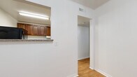 This single-floor unit offers a practical and straightforward layout with distinct zones for living, cooking, and bathing. The kitchen is equipped with rich, dark wood cabinetry extending to the upper walls, providing ample storage. Granite-patterned countertops line the workspace, complemented by a stainless steel sink, a black gas range, a microwave, and a full-size black refrigerator. Light ceramic tile flooring runs throughout the kitchen area. A pass-through window opening connects the kitchen to the adjacent hallway, enhancing the sense of openness between spaces.

The bedrooms feature dark hardwood-style flooring and are each fitted with ceiling fans with integrated lighting. One bedroom benefits from a window offering a view of surrounding greenery, bringing in natural light. The rooms maintain a clean, neutral palette with white walls and white trim throughout.

A hallway connects the main areas, transitioning between the light hardwood flooring near the kitchen and the darker flooring of the bedrooms. The bathroom is distinguished by large-format neutral tile on both the floor and walls, including a tiled walk-in shower area. Central HVAC vents are present throughout, and crown molding accents select areas of the unit.