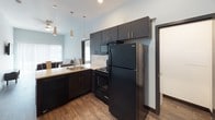 This contemporary single-floor unit is designed with a clean, modern aesthetic that carries consistently throughout every room. The open-concept living and dining area flows seamlessly into the kitchen, creating a connected and functional shared space. The living area features soaring ceilings and is furnished with a neutral sofa and accent chair, lending a comfortable yet minimalist feel. Pendant lighting above the kitchen counter adds a stylish touch to the space.

The kitchen is well-equipped with dark espresso cabinetry, a full suite of black appliances including a refrigerator, electric range, and over-the-range microwave, as well as a stainless steel sink set into light countertops. A breakfast bar with bar seating provides casual dining options.

Two bedrooms offer restful retreats, each with dark wood-style flooring, high ceilings, and windows that allow natural light to filter in. One bedroom includes a twin-sized bed frame with built-in storage, while the other accommodates a full-sized bed frame with under-bed storage drawers.

The bathroom connects directly to a walk-in closet with built-in shelving and hanging rods, offering excellent storage. Grab bars and wood-look flooring complete the practical, accessibility-conscious design. Dark door and window trim provide a cohesive contrast against the bright white walls throughout the unit.