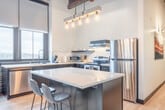 Located in the heart of Madison, WI, on East Wilson Street, this two-bedroom, one-bathroom loft seamlessly blends industrial character with modern comfort. The open-concept living and kitchen area is anchored by soaring ceilings with rich, exposed wood beams and oversized windows that flood the space with natural light and offer sweeping views of the surrounding cityscape. The kitchen is well-appointed with dark navy cabinetry, white subway tile backsplash, stainless steel appliances, a deep double sink, and a central island with seating — ideal for both everyday meals and entertaining.

Light hardwood flooring flows throughout, complementing the raw exposed brick walls that serve as a defining architectural feature. A long hallway connects the living areas to the two bedrooms, each showcasing the same exposed brick and large windows with urban views. The primary bedroom includes an in-unit stacked washer and dryer for added convenience. The bathroom is clean and contemporary, featuring marble-look tile flooring, a wood-toned vanity with a white quartz countertop, and a glass-enclosed walk-in shower. The overall ambiance is warm, stylish, and distinctly urban.