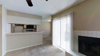 This single-floor residence offers a practical and comfortable layout with a seamless flow between the living area and kitchen. The living space is finished with plush wall-to-wall carpeting and neutral-toned walls, creating a warm and welcoming atmosphere. A ceiling fan overhead adds both functionality and a casual aesthetic to the room. The kitchen is positioned on an elevated platform and features a breakfast bar counter, overhead cabinetry, a built-in microwave, and stainless steel appliances, making it both efficient and accessible from the living area. A tiled accent detail near the sliding glass door adds a subtle architectural touch to the space. Large floor-to-ceiling sliding glass doors dressed with vertical blinds allow generous natural light to filter into the living area while providing easy access to the private balcony. The balcony, framed by a painted wood railing, offers an open view of the surrounding community, including well-maintained green lawns, a colorful children's playground, parking areas, and mature trees stretching into the surrounding neighborhood — a lively and family-friendly outdoor setting visible from the comfort of home.