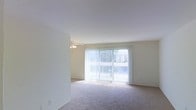 This single-floor residential space offers a clean, straightforward layout designed for comfortable everyday living. The main living area is the centerpiece of the space, featuring wall-to-wall carpeting that adds warmth and a sense of coziness underfoot. Neutral-toned walls keep the interior feeling fresh and versatile, providing an ideal backdrop for a variety of furnishing styles.

A prominent set of sliding glass doors dominates one wall of the living area, fitted with vertical blinds that allow for adjustable light control. These doors provide direct access to an outdoor area and invite generous amounts of natural light into the room, creating a bright and airy atmosphere throughout the day. The view through the glass reveals a glimpse of the surrounding outdoor environment.

A ceiling fan is centrally positioned in the living area, offering both practical air circulation and a modest decorative element. The open layout allows for flexible furniture arrangement, making the space adaptable to various living needs.

The overall ambiance is light, neutral, and move-in ready, with a simple and functional design aesthetic that prioritizes comfort and usability. The single-floor configuration ensures easy accessibility and convenient flow throughout the space.