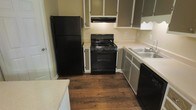 This two-story home includes a kitchen equipped with black appliances, including a full-size refrigerator, a gas range with an overhead vent hood, and a dishwasher. The kitchen features rich, dark hardwood flooring that adds warmth to the space. Cabinetry is finished in a two-tone style, with gray lower cabinets and white upper cabinets, providing generous storage throughout. Light-colored countertops offer functional prep space, and a stainless steel sink is positioned conveniently near the dishwasher. The layout follows a practical design, allowing for efficient movement between appliances and work surfaces. The neutral wall tones complement the cabinetry and flooring, contributing to a cohesive aesthetic. The home spans two floors, offering separation between living and private spaces.