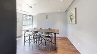 Located in the East End neighbourhood of Toronto, this single-floor communal space at 763 Woodbine Ave offers a well-appointed and functional layout designed for shared use. The interior is finished with wood-look tile flooring throughout, cool grey walls, and a clean white ceiling fitted with recessed lighting, lending the space a modern and understated aesthetic.

The dining area serves as the focal point of the room, anchored by a substantial rustic wood communal table paired with industrial-style metal stools — a combination that balances warmth with contemporary design. The seating arrangement accommodates multiple guests comfortably, making it well-suited for gatherings or shared meals.

Adjacent to the dining area is a kitchenette, outfitted with white cabinetry, a stainless steel refrigerator, microwave, dishwasher, and a sink, providing convenient amenities for food preparation and entertaining. The space is accented with small greenery, adding a touch of life to the clean, neutral palette.

A standout feature is the floor-to-ceiling glazed sliding door that opens directly onto an outdoor terrace. The terrace is furnished with seating and vibrant planters, offering views of the surrounding neighbourhood streetscape. Large windows allow abundant natural light to flood the interior, creating a bright and inviting atmosphere that seamlessly connects indoors with the outdoors.