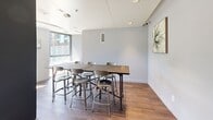 Located in the East End neighbourhood of Toronto, this single-floor communal space at 763 Woodbine Ave offers a well-appointed and functional layout designed for shared use. The interior is finished with wood-look tile flooring throughout, cool grey walls, and a clean white ceiling fitted with recessed lighting, lending the space a modern and understated aesthetic.

The dining area serves as the focal point of the room, anchored by a substantial rustic wood communal table paired with industrial-style metal stools — a combination that balances warmth with contemporary design. The seating arrangement accommodates multiple guests comfortably, making it well-suited for gatherings or shared meals.

Adjacent to the dining area is a kitchenette, outfitted with white cabinetry, a stainless steel refrigerator, microwave, dishwasher, and a sink, providing convenient amenities for food preparation and entertaining. The space is accented with small greenery, adding a touch of life to the clean, neutral palette.

A standout feature is the floor-to-ceiling glazed sliding door that opens directly onto an outdoor terrace. The terrace is furnished with seating and vibrant planters, offering views of the surrounding neighbourhood streetscape. Large windows allow abundant natural light to flood the interior, creating a bright and inviting atmosphere that seamlessly connects indoors with the outdoors.