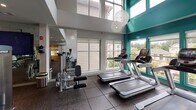 Located at 12025 Landing Place Ln in Charlotte, NC, this single-floor fitness center is a thoughtfully designed exercise facility offering a comprehensive range of equipment and an energizing atmosphere. The space is defined by its striking design elements, including soaring double-height ceilings in the cardio zone, bold teal accent walls, and expansive windows that flood the interior with natural light while offering views of the surrounding neighborhood.

The facility is divided into distinct functional zones. The cardio area houses multiple treadmills, elliptical trainers, and stationary bikes — including upright and recumbent options — along with a rowing machine, all positioned to take advantage of the generous window views. Stylish globe pendant lights hang overhead, adding a modern aesthetic touch.

The strength training zone features a multi-station cable machine, adjustable benches, and various resistance equipment, all set on durable rubber flooring. Full-length mirrors maximize the sense of space and assist with proper form. Glass partitions elegantly separate sections of the facility while maintaining an open, connected feel.

A dedicated area near the entrance displays a vibrant yellow nutrition information wall, a mounted flat-screen TV, and a textured 3D-panel accent wall, contributing to the space's contemporary and motivating design aesthetic.