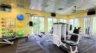 This fitness center offers a bright and welcoming environment designed to support a variety of workout routines. The space is characterized by cheerful yellow walls and large glass doors and windows that line the perimeter, flooding the interior with natural light and providing views of the surrounding greenery and landscaping outside.

The ceiling features a distinctive embossed tin-style finish, adding a decorative architectural touch, and is equipped with ceiling fans to maintain airflow during workouts. A wall-mounted television is positioned for easy viewing during exercise sessions.

The equipment layout is thoughtfully organized, with a selection of white-framed strength machines occupying the central and rear areas of the room. These include a multi-function cable machine, a leg curl/extension station, and additional resistance equipment. Rubber floor mats protect the flooring beneath the machines and free weights area.

Along one wall, a collection of stability and exercise balls in blue, yellow, and green are neatly displayed, offering options for flexibility and core training. The overall ambiance is light, airy, and functional, making this fitness center a practical and inviting space for residents or guests.