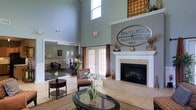 The Pavilion at Plantation Way is a beautifully appointed community clubhouse designed to offer residents a refined and welcoming gathering space. The main lounge immediately captures attention with its dramatic two-story ceilings, which allow an abundance of natural light to pour in through high clerestory windows, creating an airy and uplifting atmosphere. The focal point of the room is an elegant white-mantled fireplace set against a warm, neutral wall, adding both charm and comfort to the space. Light tile flooring throughout the main area provides a clean, polished foundation that complements the room's classic décor, which includes plush seating arrangements and tasteful accents such as floral arrangements and decorative vases. Large French doors along the perimeter invite the outdoors in and provide seamless access to exterior areas, while rich curtain panels in warm amber tones add a touch of warmth and sophistication. Adjacent to the main lounge, a secondary room visible through glass-paneled sliding doors offers additional flexible space, outfitted with its own seating and suitable for meetings or private gatherings. The overall ambiance is polished yet approachable, reflecting a community space thoughtfully designed for both relaxation and socializing.