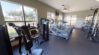 Located in Westworth Village, Texas, this single-floor fitness center offers a comprehensive workout environment designed to accommodate a wide range of exercise needs. The space is generously sized, providing ample room to move freely between equipment without feeling crowded.

The cardio section is prominently featured along the windowed walls, housing a row of Precor elliptical trainers and treadmills. Large windows line multiple walls, flooding the interior with natural light and offering pleasant views of the surrounding outdoor areas, including a deck and mature trees — an inviting backdrop during workouts.

The strength training area is equally well-appointed, featuring a multi-station cable machine, a Smith machine or functional trainer with an integrated weight stack, a dumbbell rack, barbells, weight plates, and adjustable benches. A full-length mirror spans one wall, enhancing the sense of space and allowing users to monitor their form.

Underfoot, durable rubber flooring provides safety and stability throughout. Ceiling fans are mounted overhead to help maintain air circulation. Wall-mounted televisions are positioned for visibility from the cardio equipment. The overall aesthetic is clean and functional, with neutral-toned walls and warm wood-accented trim lending a polished, welcoming atmosphere to this well-maintained fitness facility.