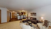This single-floor residence offers a thoughtfully designed open-concept layout that seamlessly connects the living area and kitchen, creating a cohesive and functional living space. The living room is generously sized, featuring neutral carpet flooring, soft neutral walls, and ample room for comfortable seating arrangements. Track lighting overhead complements the warm, inviting atmosphere.

The kitchen is well-equipped with rich medium-toned wood cabinetry, granite-style countertops, stainless steel appliances including a refrigerator, range, and dishwasher, and a peninsula with bar seating illuminated by pendant lighting. Wood-look flooring runs throughout the kitchen, adding warmth and contrast to the space.

A second living or flex room provides additional versatile space, finished with plush carpet, wide windows that allow natural light, and room for a variety of uses.

The bedroom is a comfortable retreat, carpeted and naturally lit through a window with blinds, featuring ample floor space and a restful ambiance.

The full bathroom includes a tub-shower combination, a vanity with dark wood cabinetry, a large mirror with overhead lighting, and durable wood-look flooring, offering both style and practicality.