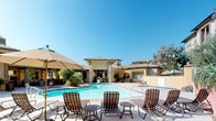 This single-story community space is designed around an expansive outdoor lifestyle, offering a well-appointed collection of amenities suited for leisure and social gatherings. At the heart of the outdoor area sits a generously sized swimming pool with crystal-clear blue water, surrounded by a wide pool deck furnished with woven strap lounge chairs and small side tables, ideal for sunbathing on clear, sunny days. An adjacent spa provides an additional relaxation option, set against a backdrop of mature trees and warm stucco architecture.

A covered terrace extends along one side of the pool area, supported by sturdy columns and outfitted with ceiling fans for comfort. This shaded lounge space features wicker-style sectional seating and armchairs, creating an inviting setting for socializing or unwinding. The architecture presents a Mediterranean-inspired aesthetic with warm tan and gold tones throughout.

Private patio areas are also available, each featuring ornate cast-iron bistro tables, woven chairs, and large shade umbrellas. One patio includes a built-in gas grill station, ideal for outdoor cooking. Lush, manicured landscaping with tropical palms, flowering shrubs, and decorative plantings borders each patio, adding color and privacy to each outdoor seating area.