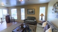This single-story home offers a comfortable and inviting atmosphere, highlighted by a well-appointed living room that serves as the heart of the space. The living area features warm, neutral-toned walls that create a relaxed and homey ambiance. Multiple windows dressed with white blinds allow generous amounts of natural light to fill the room, brightening the interior throughout the day. The space is furnished with classic, tufted upholstered seating in a muted sage green, complemented by a rich, dark wood coffee table that anchors the seating arrangement. A decorative area rug with a traditional blue and gold pattern adds texture and visual interest to the flooring. A framed landscape painting adorns the wall, serving as a tasteful focal point, while a classic table lamp with a warm shade contributes to the cozy character of the room. A ceiling fan overhead provides both comfort and subtle architectural detail. The overall design aesthetic balances traditional charm with everyday livability, making this a welcoming and functional home suited for comfortable daily living.