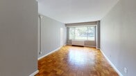 Located in the desirable Northwest Washington, DC neighborhood, Apartment 227 at 3040 Idaho Ave NW is a single-floor residence that combines classic charm with everyday functionality. The living area is a standout feature, showcasing warm, richly toned parquet wood flooring arranged in a distinctive geometric pattern that adds character throughout the space. Neutral gray walls provide a clean, versatile backdrop, while a large picture window floods the room with natural light and offers views of the surrounding greenery outside. A radiator unit beneath the window provides efficient heating. The layout flows into a separate bedroom area, also finished with the same elegant parquet flooring and neutral walls, offering a quiet and comfortable retreat. The kitchen is compact yet well-equipped, featuring warm oak cabinetry with ample upper and lower storage, a gas range, a stainless steel sink, and a full-size refrigerator set against light-toned walls. A bathroom with essential fixtures completes the unit. The overall ambiance is bright, clean, and move-in ready, making this apartment a practical and inviting option in one of Washington, DC's established residential corridors.