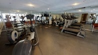 This fitness center offers a comprehensive and well-organized workout environment designed to support a variety of exercise routines. The space is outfitted with a substantial selection of cardio equipment, including treadmills, elliptical trainers, and stationary cycles, arranged to maximize accessibility and flow throughout the room. Durable rubber flooring covers the entire area, providing both safety and comfort underfoot during high-intensity workouts. Overhead, ceiling-mounted flat-screen televisions are positioned for easy viewing from multiple workout stations, adding an entertainment element to training sessions. Wall-to-wall mirrors line the perimeter, visually expanding the space and allowing users to monitor their form while exercising. Recessed lighting in the textured ceiling provides consistent, even illumination throughout. The overall layout is thoughtfully designed to accommodate multiple users simultaneously, with ample spacing between machines to ensure a comfortable and efficient workout experience. The clean, functional aesthetic of the space reflects a focus on performance and usability, making it a practical and well-appointed facility for fitness enthusiasts of all levels.