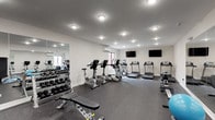 The fitness center at The Lofts at Middlesex is a spacious, single-floor facility designed to support a comprehensive workout routine. The room features a clean, modern aesthetic with bright white walls, recessed lighting, and durable rubber flooring throughout, creating a functional and inviting environment for exercise.

The cardio section is well-stocked with multiple True-brand treadmills, elliptical trainers, and stationary bikes, arranged to allow comfortable spacing between equipment. Two wall-mounted flat-screen televisions are positioned to keep users entertained during cardio sessions. A dedicated wall-unit air conditioner and a window offering views of the surrounding building exterior ensure the space remains comfortable during use.

The strength training area includes a multi-station cable and weight machine, an adjustable weight bench, and a full rack of dumbbells in varying sizes. A rowing machine adds additional versatility to the cardio and strength options available. Stability balls and BOSU balance trainers are also on hand for functional fitness training.

Large mirrors line sections of the walls, visually expanding the space while allowing users to monitor their form. The combination of recessed ceiling lights and flush-mount fixtures provides even, well-distributed illumination throughout the entire fitness center, maintaining a bright and energetic atmosphere.