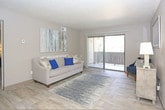 Located at 5222 Wyoming Blvd NE in Albuquerque, New Mexico, this single-floor residence offers a thoughtfully designed layout with comfortable living spaces throughout. The living room is a welcoming area featuring wood-style flooring, neutral-toned walls, and a large sliding glass door that opens to a private balcony, flooding the space with natural light and providing outdoor views. A dedicated dining area sits nearby, complete with a ceiling fan and a decorative round mirror, creating an intimate setting for meals. The kitchen is equipped with white cabinetry, granite-style countertops, a stainless steel sink, and black appliances, offering a clean and functional workspace. The bedroom is generously sized with plush carpeting, a textured ceiling, and a window dressed with vertical blinds that allow for adjustable light. It connects conveniently to a well-appointed bathroom featuring wood-style flooring, dual-tone walls in gray and blue, grab bar-equipped shower with a decorative curtain, and tasteful botanical artwork. The overall ambiance of the residence is calm and cohesive, with a neutral color palette accented by cool blue tones carried throughout the décor, creating a comfortable and visually harmonious living environment.