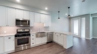 This well-appointed single-floor residence showcases a thoughtfully designed interior with a cohesive modern aesthetic. The heart of the home is the open-concept kitchen, which is outfitted with a full suite of stainless steel appliances, including a gas range, over-the-range microwave, dishwasher, and a French door refrigerator. Crisp white shaker-style cabinetry extends to the upper walls, complemented by a classic subway tile backsplash and stone countertops with natural veining. A stainless steel farmhouse sink adds a refined focal point, while two industrial-style pendant lights hang overhead, providing warm, focused illumination. The kitchen flows seamlessly into the adjacent living area, anchored by durable hardwood-style plank flooring that runs throughout the space. The bedroom is a calm, neutral retreat featuring large windows that invite abundant natural light and a sliding glass-panel door, adding an elegant architectural detail. The bathroom is cleanly finished with stone-topped vanity cabinetry, coordinating hardwood-style flooring, and a walk-in shower equipped with a grab bar, reflecting both style and accessibility. Recessed lighting and a textured ceiling are consistent throughout, lending the entire residence a polished, move-in-ready appeal.