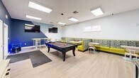 Located at 304 Madison Ave in Boone, NC, this single-floor game and lounge space is thoughtfully designed for recreation and social interaction. The room makes a strong visual statement with its bold two-tone color palette — a deep navy blue accent wall contrasts with lighter blue-gray and crisp white walls — creating an energetic yet inviting atmosphere throughout.

At the heart of the space sits a full-size pool table positioned beneath a striking geometric pendant light fixture, serving as the room's focal point. Recessed and flush-panel ceiling lights ensure the area is well-lit, complemented by a skylight that introduces natural daylight. Light-toned wood-style flooring runs throughout, adding warmth and continuity.

Along one wall, a generous L-shaped booth with vibrant yellow-green upholstery and patterned backrests provides ample lounge seating, accompanied by small café-style tables ideal for games or conversation. High-top bar tables and stools occupy another corner, offering an alternative social setting. Two wall-mounted flat-screen TVs provide additional entertainment options.

The deep blue wall houses a practical credenza with storage, a cue rack, and a whiteboard, keeping the space organized. A glass-panel door and a standard entry door provide access and allow natural light to filter in, rounding out this well-appointed recreational environment.