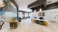 The Narcoossee Cove leasing office is a thoughtfully designed, single-floor space that balances professional functionality with sophisticated interior styling. Visitors are welcomed through a grand entry foyer with soaring ceilings, large transom-topped glass doors, and light travertine tile flooring that continues throughout the entire space, creating a seamless and airy flow.

The centerpiece of the lobby is a dramatic floor-to-ceiling abstract mural, rendered in rich earth tones and deep blues, anchoring a cozy seating vignette furnished with plush olive-green velvet armchairs and a warm wood accent table. This artistic focal point sets a refined tone that carries throughout the space.

The reception area features a curved, wood-paneled front desk with a crisp white countertop, complemented by elegant gold pendant lights suspended from a boldly contrasting dark ceiling soffit above. Shiplap accent walls add texture and warmth behind the desk.

Toward the rear, a relaxed lounge area offers tufted black leather wingback chairs arranged beside large floor-to-ceiling windows that frame lush outdoor views of palm trees, a pool deck, and surrounding greenery. A conference-style dining table nearby provides additional functionality, bathed in natural light from expansive windows. Framed abstract artwork punctuates the walls throughout, reinforcing the space's upscale, contemporary character.