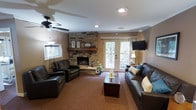 This single-floor clubhouse offers a warm and inviting gathering space designed for comfort and community. The main lounge area serves as the centerpiece of the clubhouse, anchored by a striking floor-to-ceiling stacked stone fireplace with a rustic wooden mantel, providing a cozy focal point for the room. The space is furnished with rich, dark leather sofas and armchairs arranged around a wooden coffee table, creating an ideal setting for relaxation and socializing. A wall-mounted flat-screen television adds to the entertainment options available to guests. Natural light flows generously into the room through a set of classic French doors, which open to an adjacent outdoor area, as well as a side window dressed with horizontal blinds. The ceiling features a ceiling fan with integrated lighting and recessed can lighting, ensuring the space is well-lit and comfortable throughout the day. Warm, neutral-toned walls and carpeted flooring contribute to the overall inviting ambiance. Decorative accents, including lanterns on the fireplace mantel and accent pillows in teal and yellow, add subtle pops of color and personality to the space, making this clubhouse a well-appointed retreat for its community.