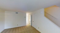 This two-story townhome features a clean, neutral interior designed for comfortable everyday living. The main living area is finished with wall-to-wall carpeting and bright white walls, creating a light and airy atmosphere. Crown molding adds a classic architectural touch, while a centrally located HVAC vent ensures climate control throughout the space. A staircase connects the two floors, with warm yellow accent walls lending a welcoming feel to the transition between levels. An adjacent hallway, illuminated by a small chandelier, leads to additional rooms, hinting at a thoughtful and connected floor plan. A closet door near the staircase provides convenient built-in storage on the main level. The overall layout is open and uncluttered, making it easy to personalize and furnish to taste. With two bedrooms and two full bathrooms distributed across both floors, the townhome offers a practical separation of living and private spaces. The neutral color palette and classic detailing throughout give the home a timeless, move-in-ready appeal suited for a variety of lifestyles.