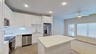 This single-floor unit offers a clean, contemporary design with a cohesive neutral color palette and light hardwood-style flooring throughout the main living areas. The open-concept kitchen is a standout feature, showcasing crisp white shaker cabinetry that extends to the ceiling, a classic white subway tile backsplash, and bright white quartz countertops. A central island provides additional prep space and storage. Stainless steel appliances — including a French door refrigerator with ice and water dispenser, dishwasher, range, and over-the-range microwave — complete the kitchen. Recessed lighting keeps the space bright and functional. The kitchen flows naturally into the dining area and living room, where a ceiling fan, large windows, and a sliding glass door fill the space with natural light and provide outdoor access. The bedroom area is generously sized with a large window and door accessing the outdoors. The bathroom features neutral tile flooring, white shaker-style cabinetry, a backlit vanity mirror, and a soaking tub, with direct access to a walk-in closet. Throughout, the design emphasizes clean lines, modern finishes, and a light, airy atmosphere.