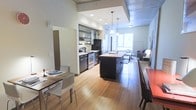 This single-floor space showcases an open-concept design that seamlessly connects the kitchen, dining, and living areas into one cohesive environment. The kitchen is outfitted with dark cabinetry, stainless steel and black appliances, a range with hood vent, and generous counter space along the perimeter. A central island with a butcher-block-style countertop provides additional prep space and doubles as a casual dining surface, complemented by modern bar-height seating. Pendant lighting hangs above the island, adding both task illumination and visual interest. The dining area accommodates a full table with seating for multiple guests and is positioned to benefit from natural light filtering in through the windows at the far end of the space. The living area, visible in the background, offers a comfortable and relaxed setting. Throughout, warm-toned hardwood flooring unifies each zone and enhances the overall brightness of the interior. The ceiling transitions between smooth white surfaces and an exposed concrete finish, lending an industrial-modern character. The layout is efficient yet open, creating a welcoming and functional living environment.