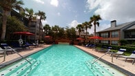 This outdoor amenity space is designed to offer a well-rounded recreational experience centered around a generously sized swimming pool with clear turquoise water. The pool is framed by a wide, tiled sundeck furnished with navy blue lounge chairs, providing ample space for sunbathing under open skies. Tall palm trees and mature shade trees are interspersed throughout the area, adding a tropical ambiance and natural canopy. Red market umbrellas positioned around the deck offer additional shade and visual interest.

Adjacent to the pool is a covered outdoor lounge area outfitted with a ceiling fan and open-air openings on multiple sides, allowing for comfortable seating with direct sightlines to the pool. Navy blue cushioned sectional seating with accent pillows creates a relaxed gathering space ideal for socializing. The surrounding multi-story buildings feature brick and siding exteriors with large windows and covered balconies, enclosing the pool area in a courtyard-like setting.

Access to the pool area is managed through iron-railed gated entries, while tile-floored walkways connect the covered lounge to the broader deck. Manicured hedgerows and planted greenery contribute to a polished, resort-inspired atmosphere throughout the space.