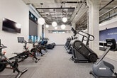 Located at 4651 Dodge St in Omaha, Nebraska, this contemporary fitness facility delivers a comprehensive wellness experience within a thoughtfully designed, single-floor space. The industrial-inspired aesthetic is defined by exposed concrete ceilings with visible ductwork and piping, polished concrete floors, and bold black steel accents throughout — creating a modern, urban atmosphere.

The main exercise floor is expansive and well-equipped, housing a full array of cardio machines including treadmills, StairMasters, stationary bikes, and ellipticals, alongside strength and resistance training equipment. Oversized globe pendant lights illuminate the space warmly, complementing the natural light that filters in through a band of windows along the upper walls. A motivational "Push Your Horizon" feature wall with built-in storage adds a branded focal point.

A dedicated studio room with warm hardwood flooring, floor-to-ceiling mirrors, and large glass walls provides an ideal space for group classes or personal training. An adjoining room features built-in locker-style storage and a mobile presentation board. A dramatic custom steel staircase with geometric patterned railings serves as a striking architectural detail. The facility also includes a cedar-lined infrared sauna with ambient LED lighting, offering a premium recovery experience. Glass-walled partitions and exterior access doors with outdoor views complete this polished, full-service fitness environment.