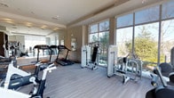 This fitness center offers a clean, contemporary workout environment designed to support a full range of exercise routines. The space is anchored by large, floor-to-ceiling windows that flood the room with natural light and provide refreshing views of the surrounding greenery, creating an inviting atmosphere for physical activity. The neutral color palette — warm beige walls, light-toned ceilings, and grey carpet flooring — contributes to a calm, focused ambiance throughout. A full wall of mirrors along one side visually expands the space while allowing users to monitor their form during workouts. The equipment selection is modern and well-maintained, featuring multiple treadmills for cardio training alongside a variety of professional-grade strength and resistance machines. The layout is thoughtfully organized, providing ample space between equipment for comfortable and safe use. Recessed lighting complements the natural light, ensuring the space remains well-lit at all times. The overall design balances functionality with an aesthetic sensibility, making this fitness center a practical and pleasant destination for daily exercise.