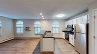 This single-floor unit offers a clean, functional layout with a cohesive design carried throughout each room. The open-concept kitchen and living area serves as the heart of the space, anchored by a central island with dark granite countertops and a built-in sink. White shaker-style cabinetry lines the kitchen walls, complemented by stainless steel appliances including a refrigerator, microwave, and electric range. Light hardwood-style flooring runs throughout the main living areas, adding warmth and continuity.

A signature architectural detail is the series of arched windows found across multiple rooms, allowing generous natural light to fill the interior and creating an airy, open ambiance. The open living area adjacent to the kitchen provides ample space and features recessed lighting and track lighting, offering flexible illumination options. Baseboard heating is present throughout the unit.

The bedrooms are carpeted for comfort, with one featuring a ceiling fan and a pair of arched windows. Additional bedrooms are well-sized with neutral tones and baseboard heating. The bathroom is equipped with a full soaking tub and shower combination, grab bars for accessibility, and the same light hardwood-style flooring found in the main areas, completing this thoughtfully appointed space.